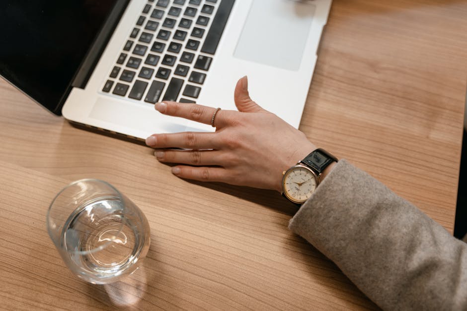 A close-up of a woman's watch-adorned hand on a wooden desk beside a laptop and glass of water.